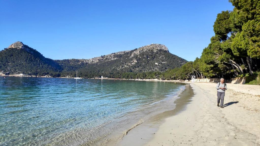 Hombre caminando por la playa de Formentor, con aguas cristalinas y montañas al fondo bajo un cielo azul.