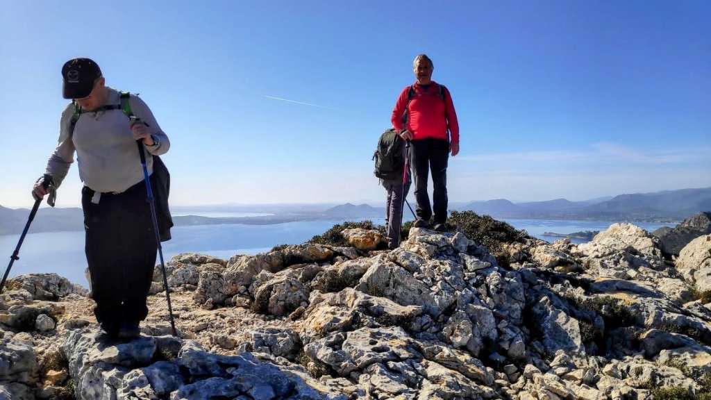 Grupo de senderistas en la cima del monte Na Blanca, con vistas panorámicas al mar y la costa. Uno de ellos se agacha usando bastones de trekking, mientras otro posa de pie con una mochila roja.