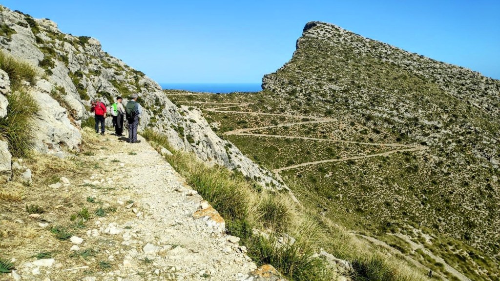 Grupo de senderistas caminando por el Coll de la Bretxa en un paisaje montañoso, con vistas al mar y un pico prominente al fondo.