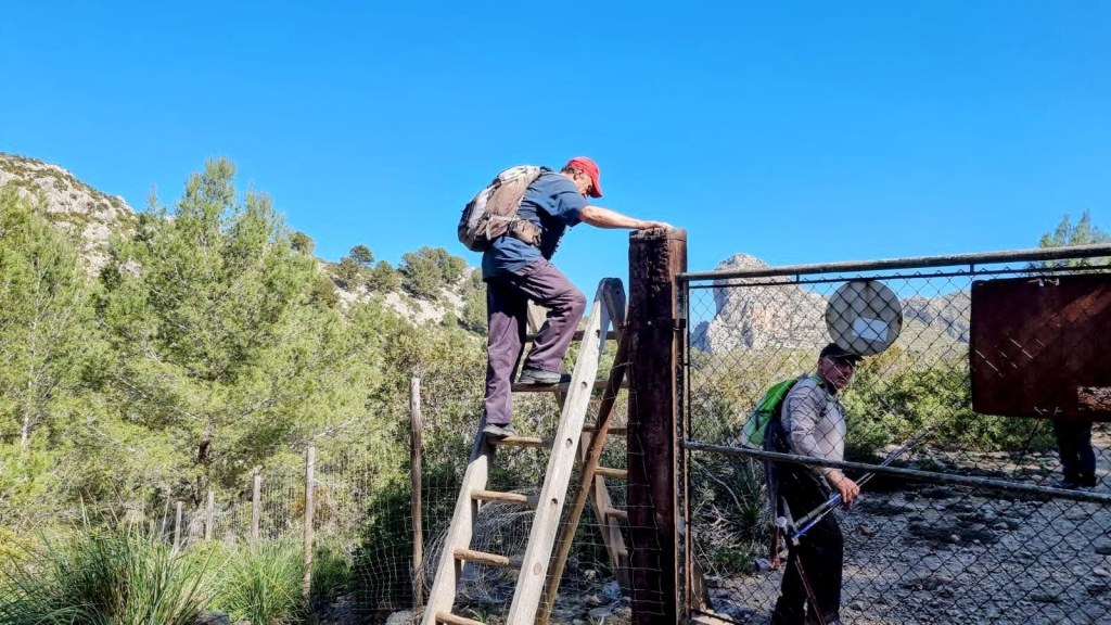 Persona subiendo por una escalera, mientras otra persona permanece detrás cerca de una puerta de metal en un entorno natural con vegetación y montañas al fondo.