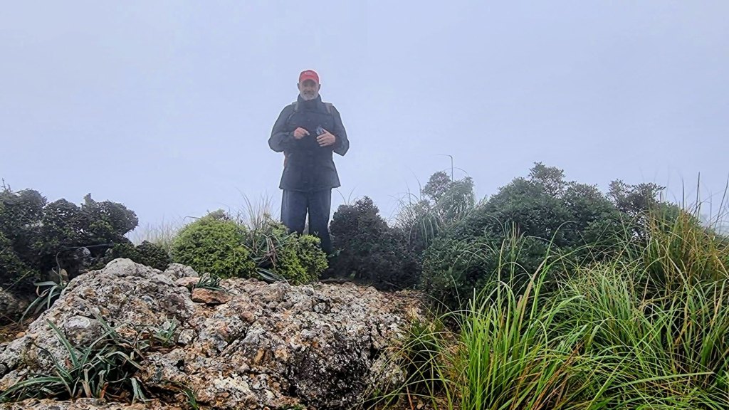 Hombre de pie en la cima del Puig de Sant Miquel, rodeado de niebla y vegetación, mientras sostiene una cámara.