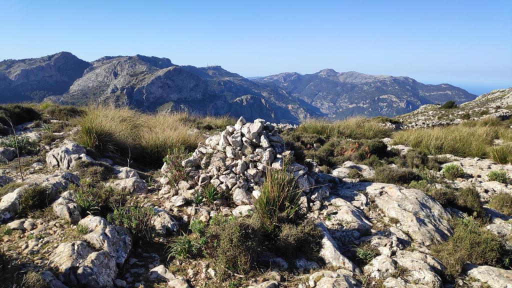 Vista panorámica desde el Coll de ses Solanes, con un montón de piedras y vegetación en primer plano y montañas al fondo.