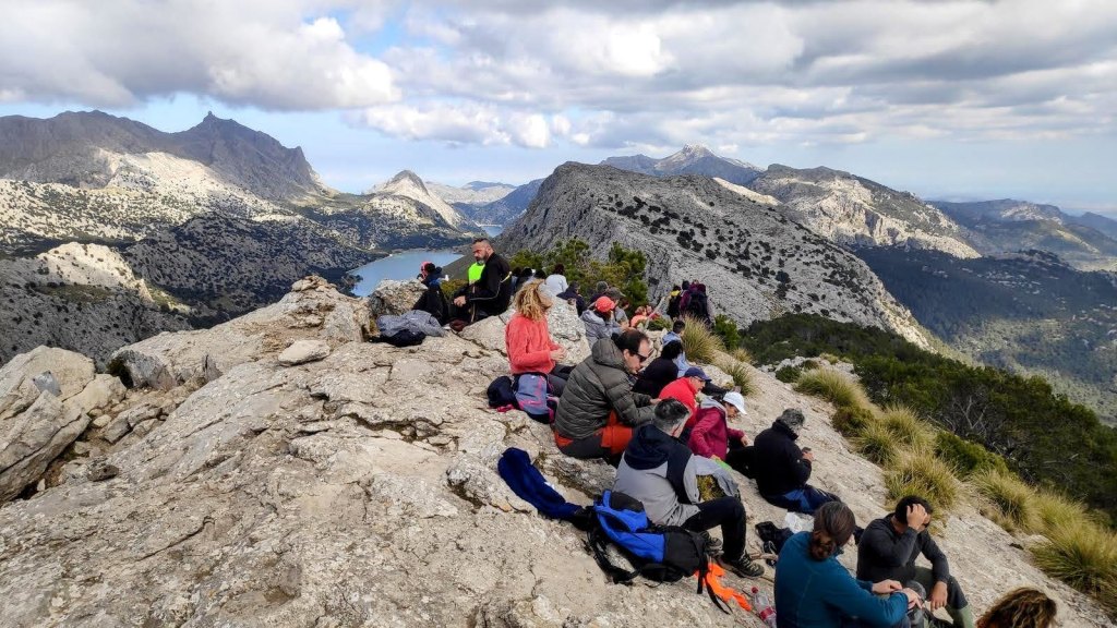 Grupo de excursionistas sentados en una roca en la cima del Puig de l’Ofre, con vistas panorámicas del paisaje montañoso y un embalse al fondo.