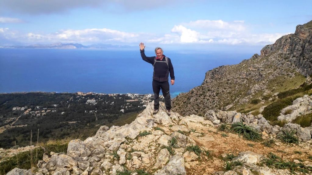 Hombre de pie en el Puig del Migdia,  levantando la mano, con un paisaje costero y montañoso al fondo.