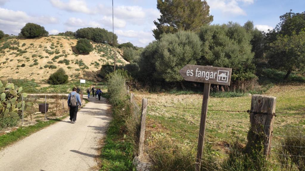 Ruta de senderismo con señalización hacia 'es fangar', rodeada de campos rurales y colinas en un día nublado.