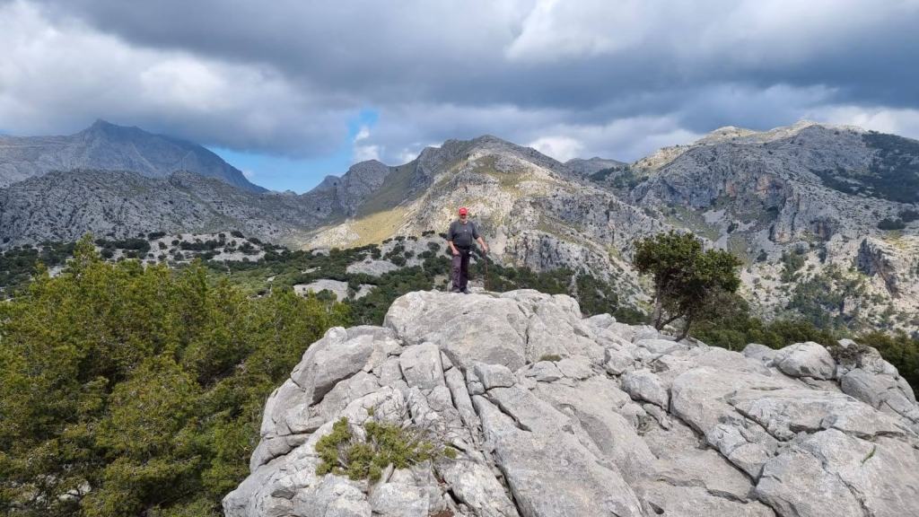 Persona de pie en el Puig de sa Cova dels Carboners con una vista panorámica de montañas y nubes oscuras de fondo.