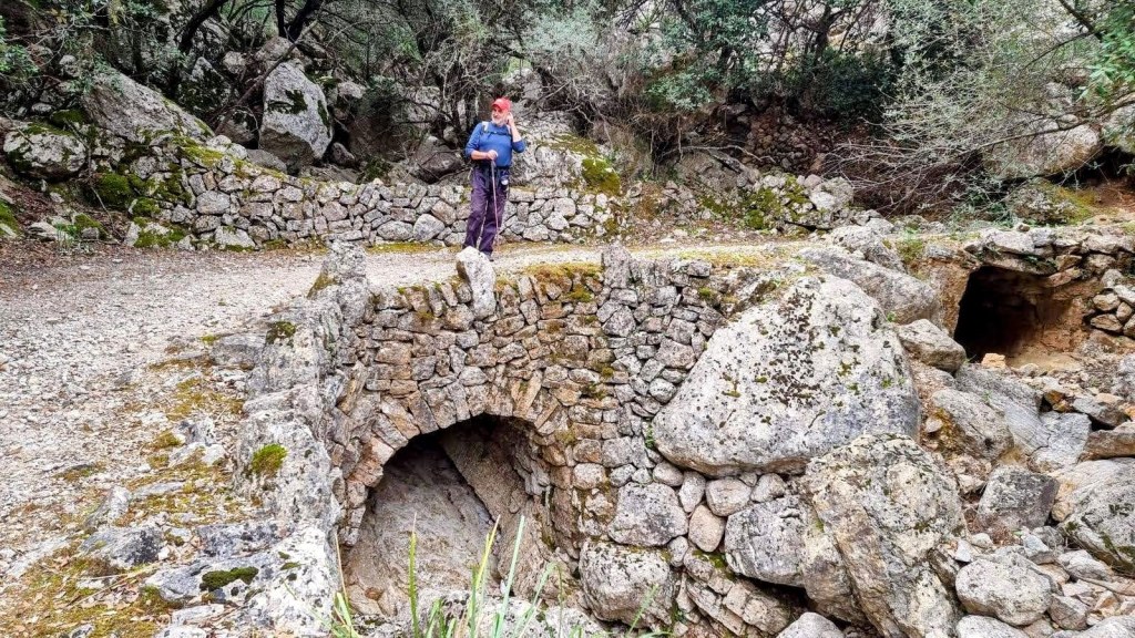 Puente de piedra en un sendero rodeado de vegetación en la ruta 'Puig d'Alfàbia por el Pas de Llis', con una persona de pie observando.