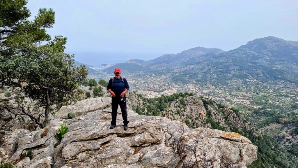 Hombre de pie sobre una roca, con fondo montañoso y vista panorámica del valle de Sóller y el mar al fondo.
