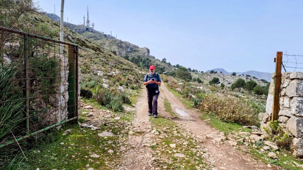 Senderista caminando por un camino de tierra en la Sierra de Alfàbia, con antenas visibles en la parte superior de la montaña y un paisaje natural en el fondo.