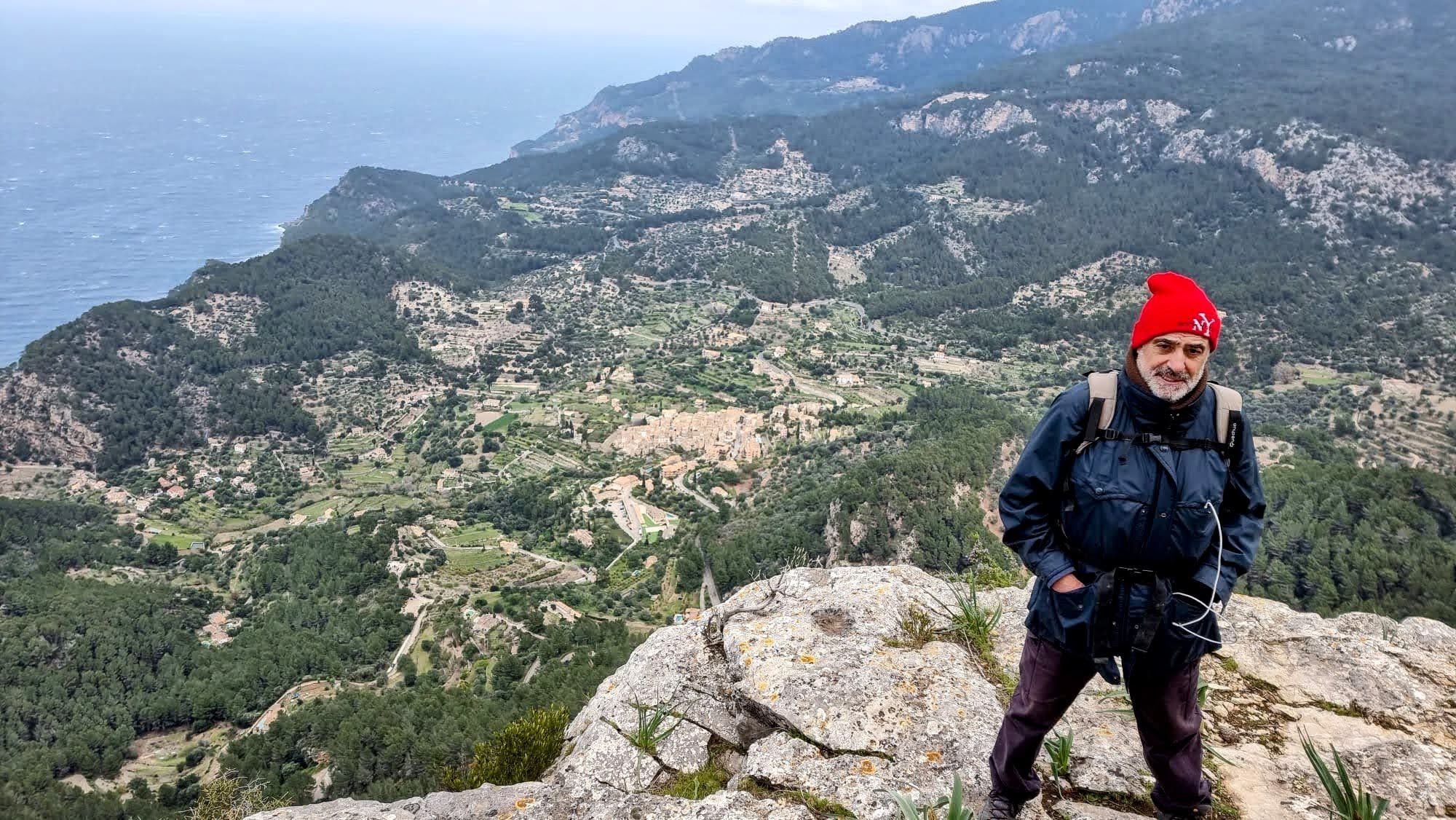 Hombre de pie en la cima del Morro de ses Serveres, con vista panorámica del paisaje montañoso y el mar al fondo, usando un abrigo y un gorro rojo.