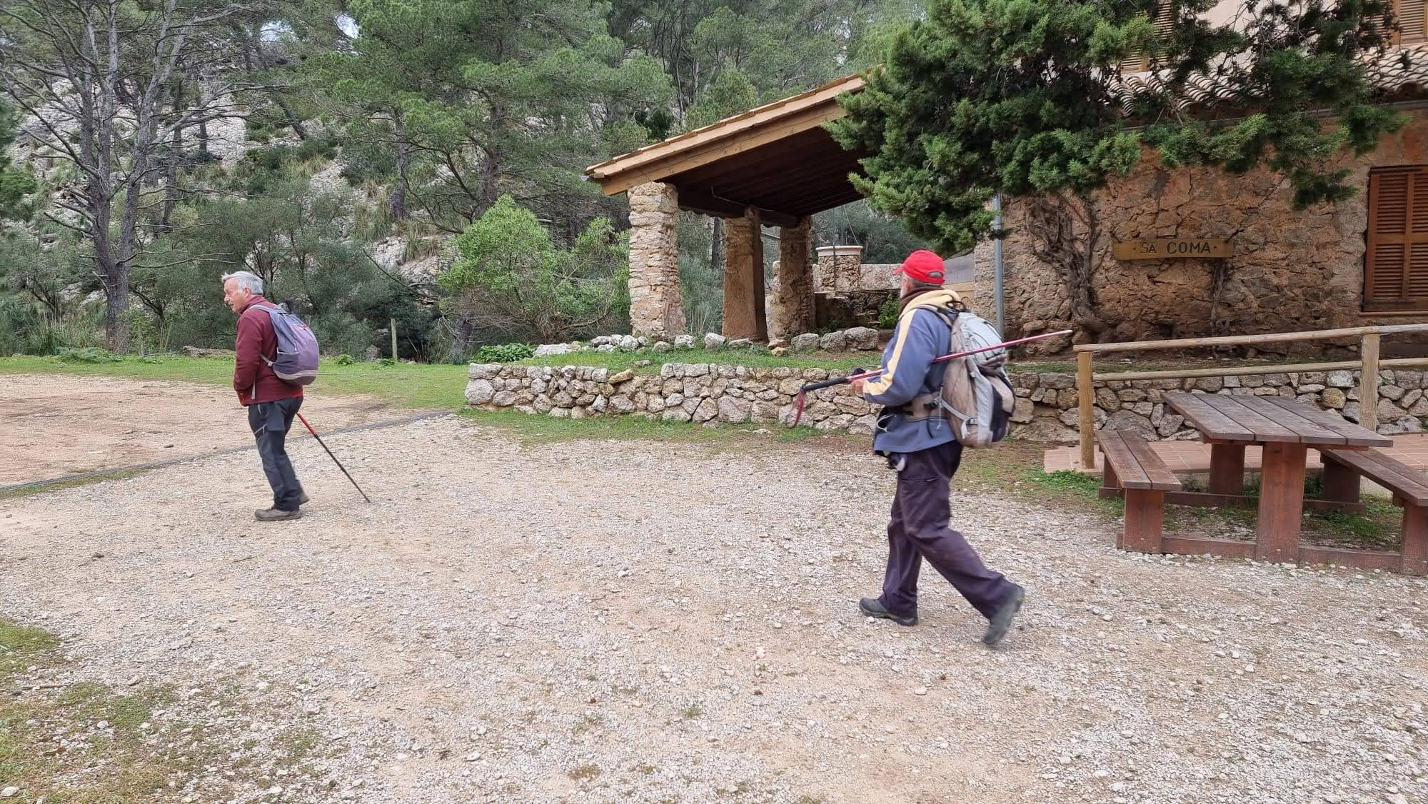 Serra des Pinotells desde Estellencs - Caminando por Mallorca Dos excursionistas caminando por un camino de tierra junto al Refugio de la Coma d’en Vidal, en la naturaleza, con mesas de picnic al costado y árboles alrededor.