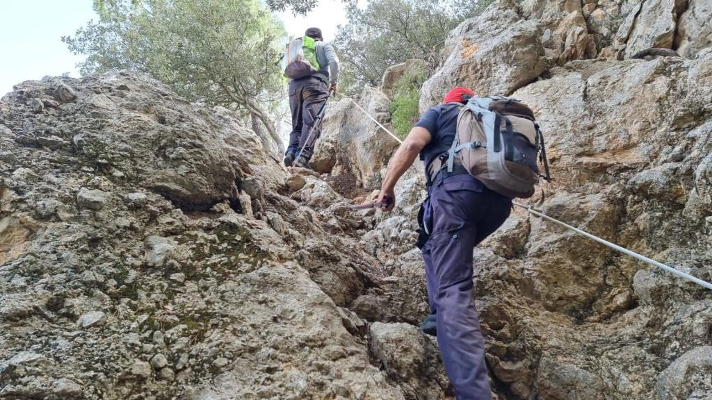 Dos excursionistas ascendiendo por un sendero rocoso, ayudándose de una cuerda de seguridad en un terreno irregular y montañoso.