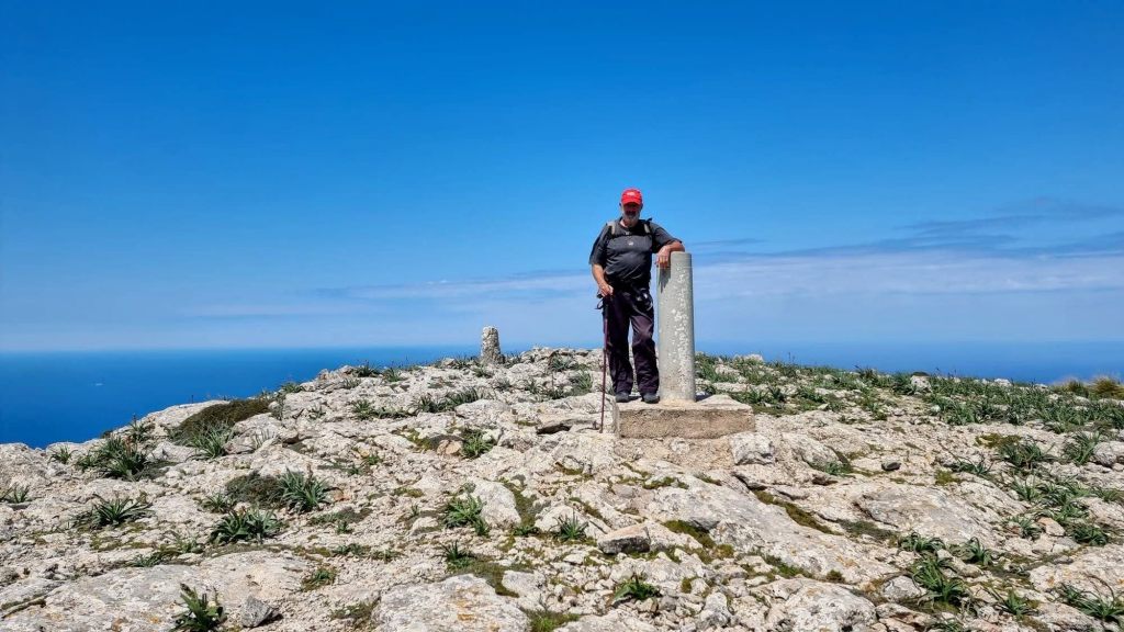 Hombre de pie en la cima de la Mola de s'Esclop, junto a un vértice geodésico, con un paisaje montañoso y un cielo azul de fondo.