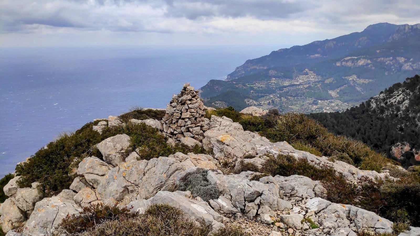 Serra des Pinotells desde Estellencs - Caminando por Mallorca Vista panorámica desde la cima de Es Morralàs, con un mojón de piedras en primer plano, rodeado de rocas y vegetación, y el mar en el fondo.