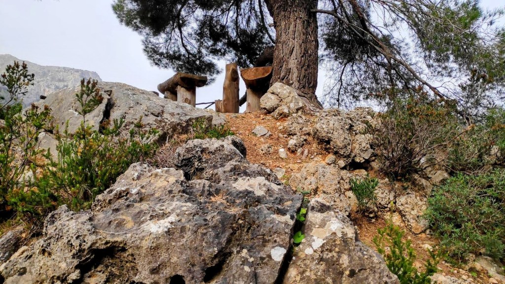 Mirador natural con mesas y sillas de troncos de madera, rodeado de rocas y vegetación arbustiva, en un entorno montañoso.