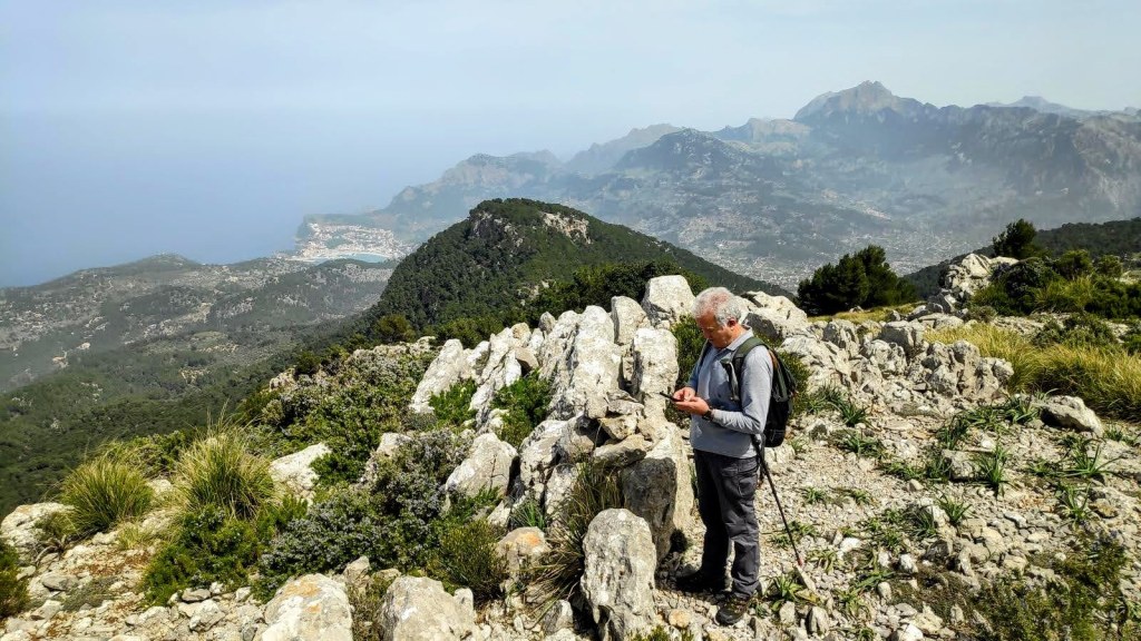 Un hombre se encuentra de pie en la cima del Puig de Sa Galera, observando su teléfono, rodeado de rocas y vegetación. En el fondo, se puede ver un paisaje con montañas y el mar.