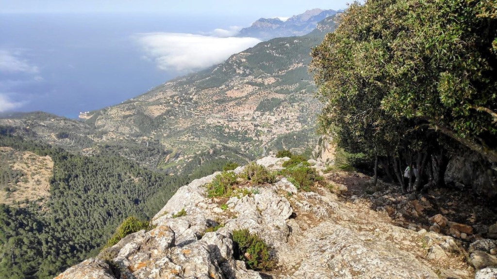 Vista panorámica desde un peñasco, mostrando un paisaje montañoso con vegetación, caminos y una vista parcial del mar en la distancia.