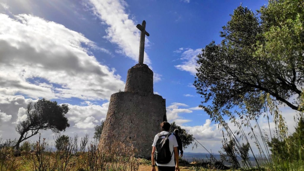 Persona caminando hacia la Cruz de Minyó en el Puig de Santa Magdalena, rodeada de vegetación y un cielo parcialmente nublado.