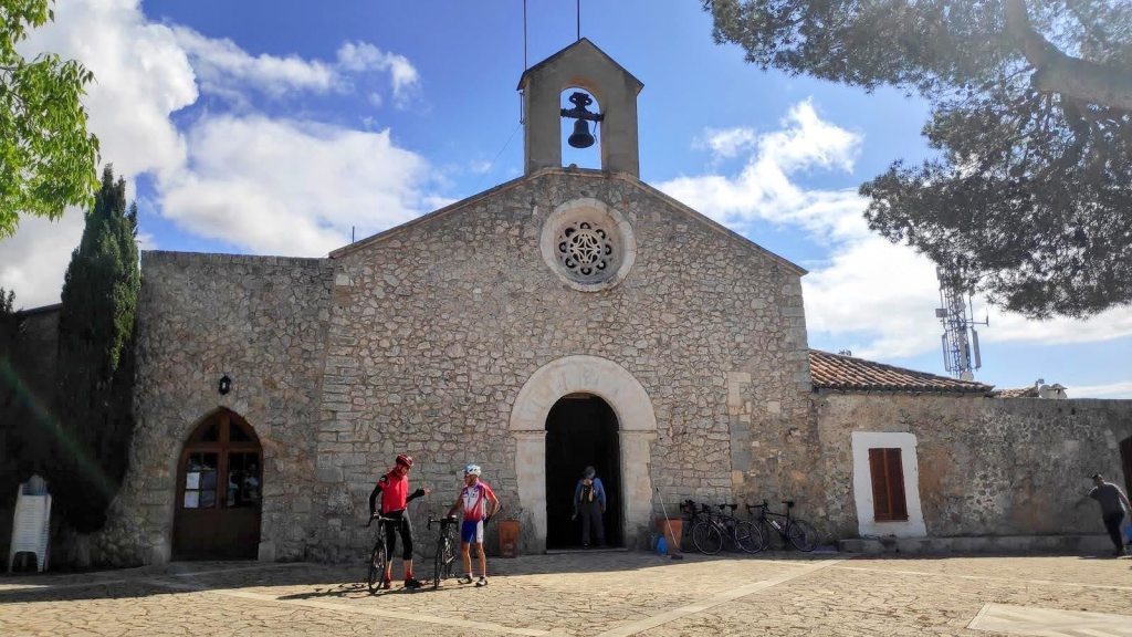Fachada de la ermita de Santa Magdalena, con detalles arquitectónicos notables y visitantes junto a bicicletas. Cielo parcialmente nublado.
