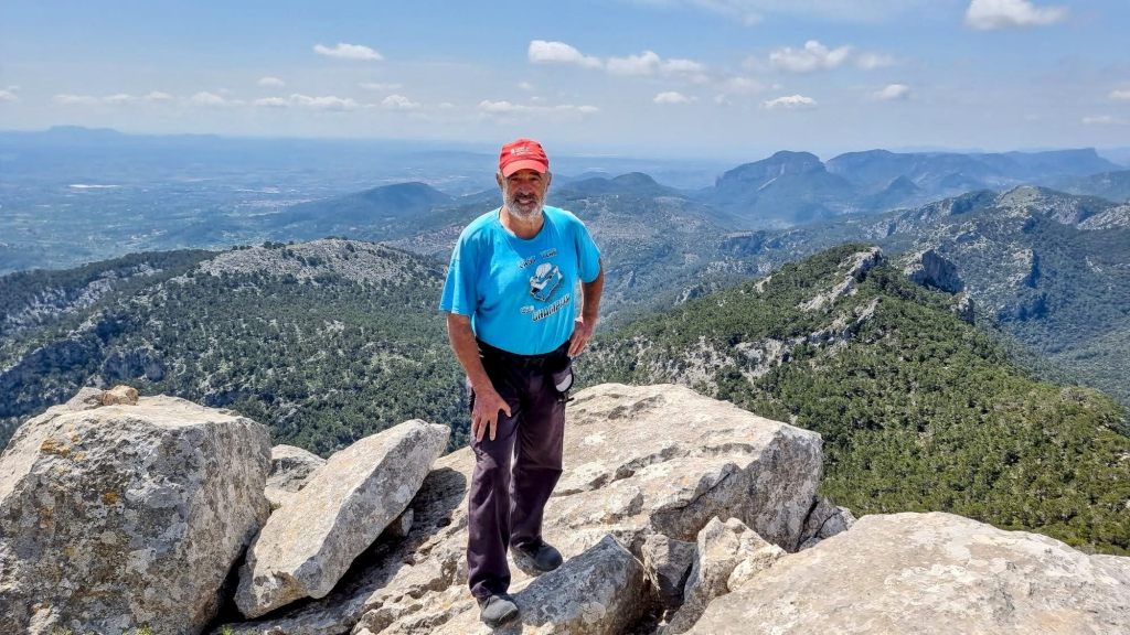 Un hombre de pie en la cima del Puig de n'Alí, rodeado de rocas y un paisaje montañoso que se extiende al fondo bajo un cielo claro.