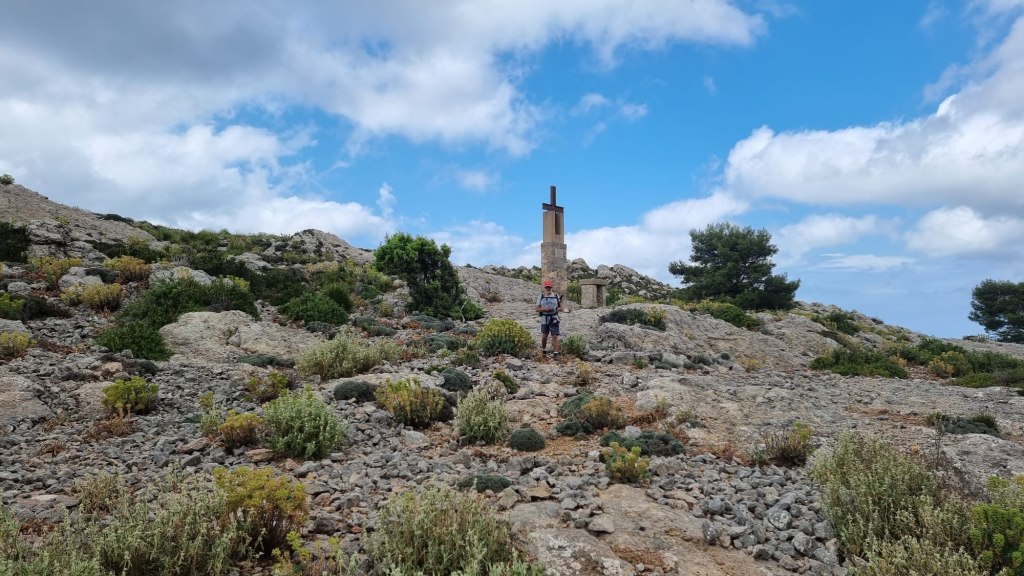 Vista de un senderista en el Coll des Cards Colers, rodeado de vegetación y rocas, bajo un cielo nublado.