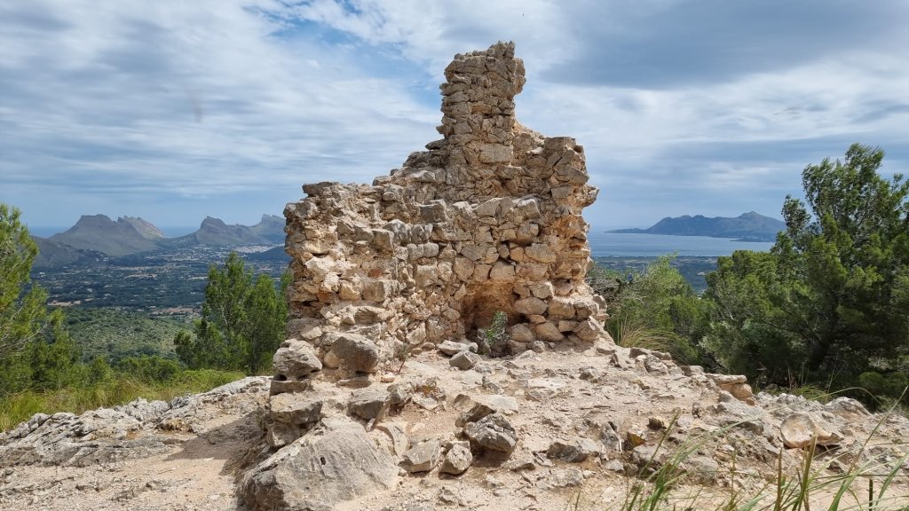 Ruinas de una estructura de piedra con vistas panorámicas hacia la costa y montañas en el fondo, bajo un cielo nublado.