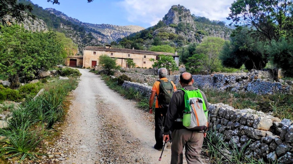 Dos excursionistas caminando por un camino de tierra rodeado de vegetación, con  Can Bajocaal fondo en las montañas de Serra de Tramuntana.