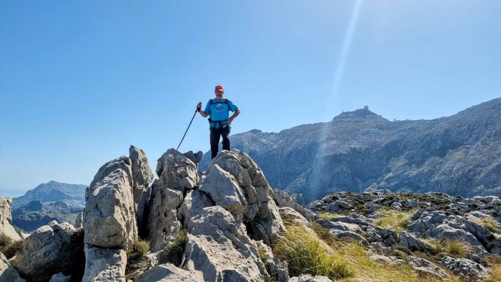 Hombre de pie sobre una roca en la cima del Puig de s’Alzinar sosteniendo un bastón, con un paisaje montañoso y un cielo despejado de fondo.