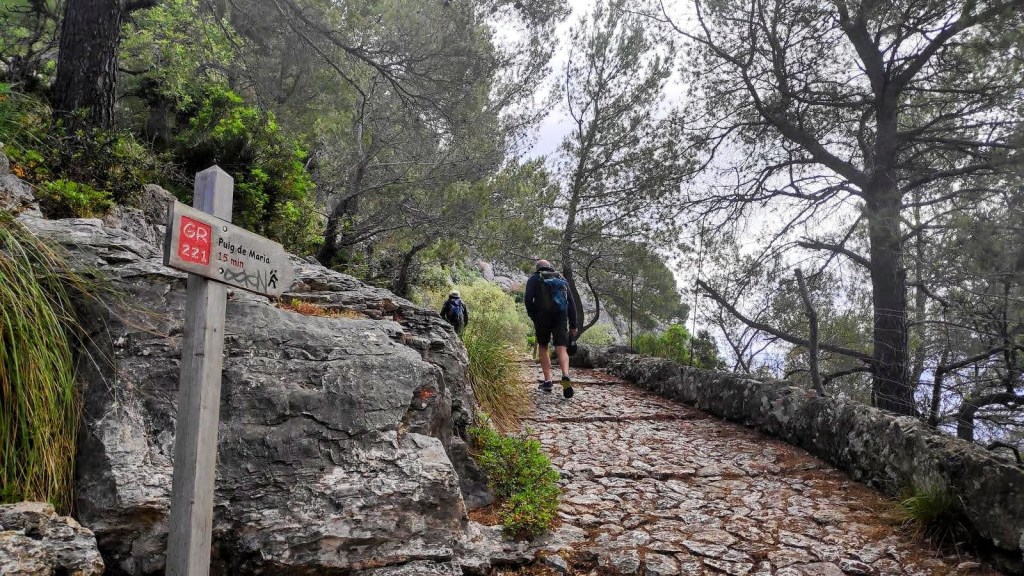 Sendero empedrado del GR-221 hacia el Puig de María, con un caminante y señalización indicando la dirección.