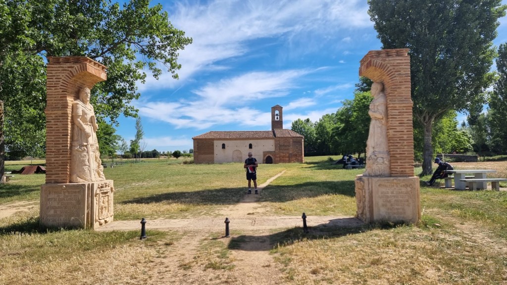 Etapa 18:Ledigos-Bercianos del Real Camino - Camino de Santiago Escultura de peregrinos en un camino con la Ermita de la Virgen del Puente al fondo y un cielo azul despejado.