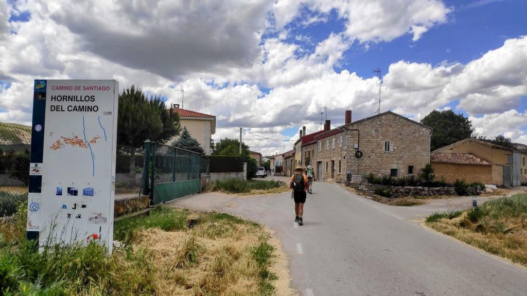 Imagen que muestra un cartel del Camino de Santiago en Hornillos del Camino, con un peregrino caminando por una carretera rural flanqueada por casas y un cielo nublado.
