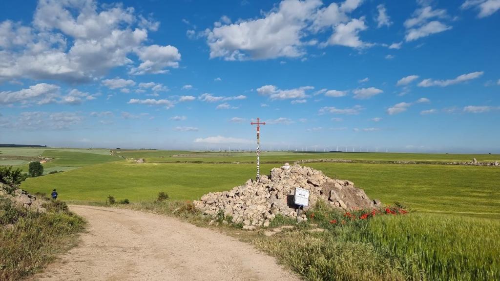 Vista del campo con extensas áreas de cereales y la Cruz de Santiago sobre un montón de piedras, bajo un cielo azul con nubes blancas.