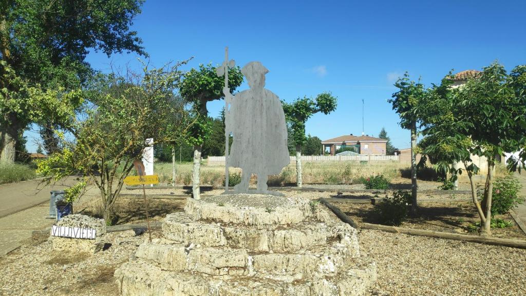 Monumento al peregrino en Villovieco, representando a un caminante con un bastón, rodeado de vegetación y señales del camino.