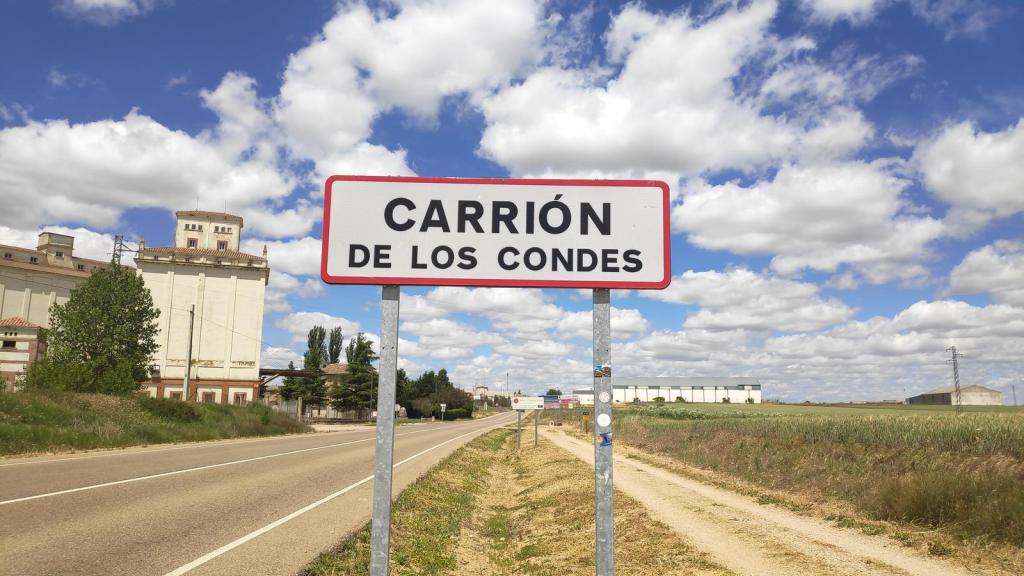 Cartel que señala la entrada a Carrión de los Condes, con un cielo nublado y paisaje rural alrededor.