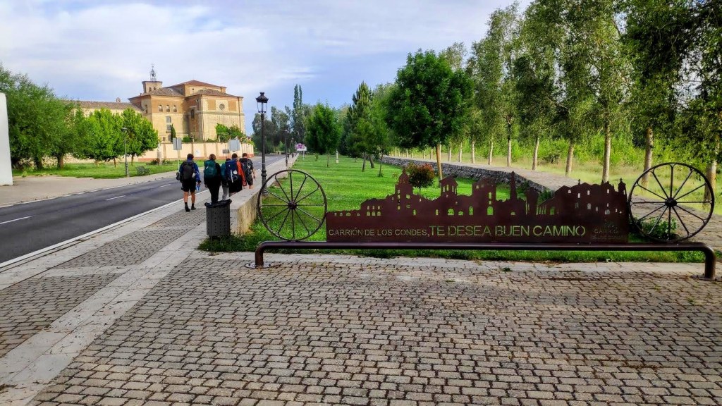 Vista de un camino en Carrión de los Condes con un cartel que dice 'Carrión de los Condes, te desea buen camino' y un fondo de árboles y edificios.