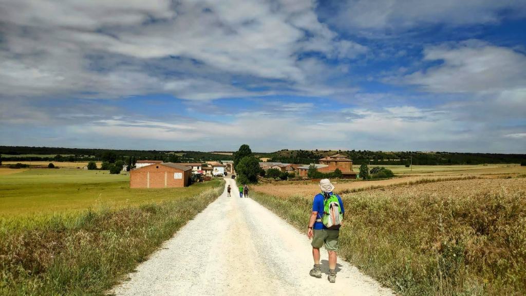 Un grupo de peregrinos camina por un camino rural hacia una pequeña localidad, rodeados de campos de cultivo y bajo un cielo despejado.