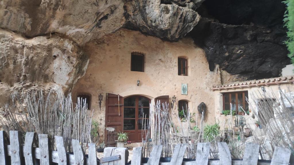 Vista de una casa construida bajo una gran roca en un paisaje rural, con ventanas y puertas de madera, decoraciones y un cercado de madera en primer plano.
