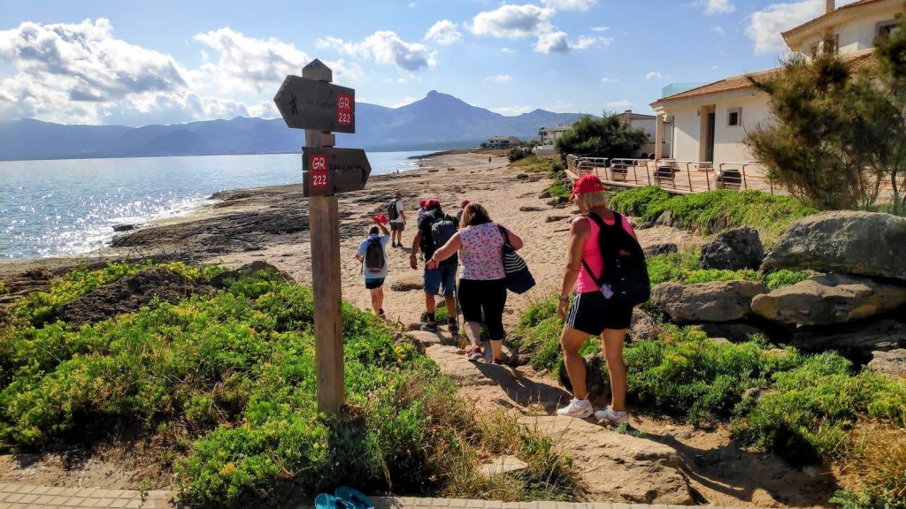 Grupo de personas caminando por el sendero costero que lleva a la Colònia de Sant Pere, con señalización del GR-222 y panorámica del mar y montañas al fondo.