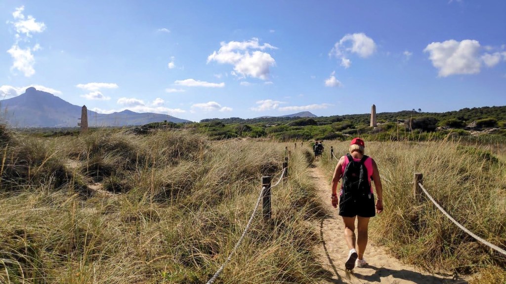 Persona caminando por un sendero de arena rodeado de hierba alta, con montañas al fondo bajo un cielo parcialmente nublado.