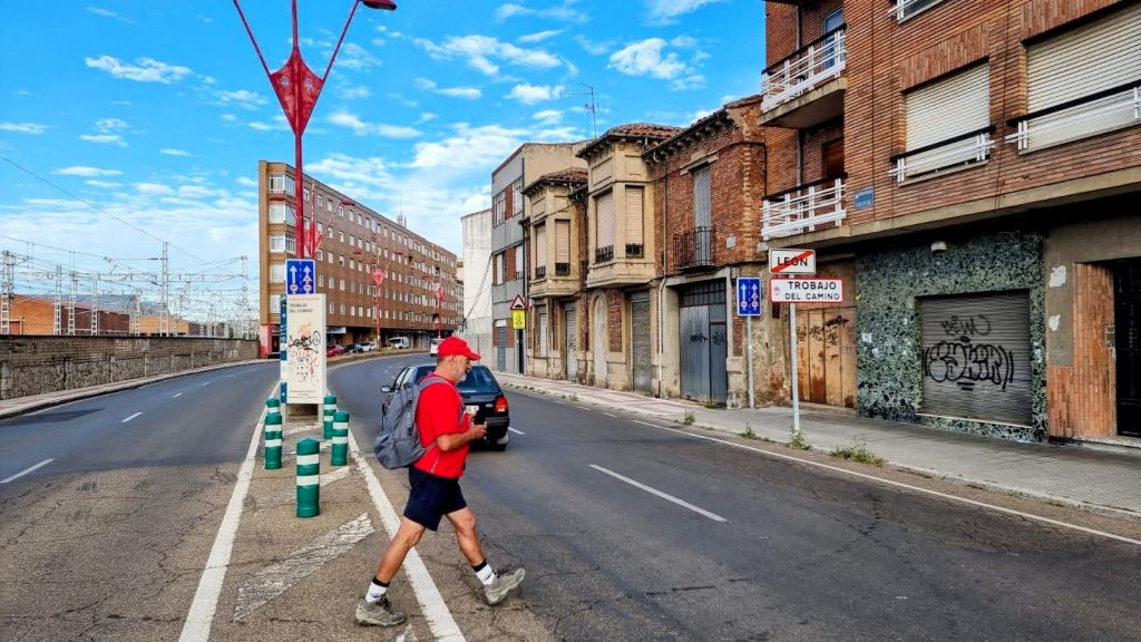 Un peregrino camina por la avenida en dirección a Trobajo del Camino, con edificios y señales de tránsito alrededor, bajo un cielo azul.