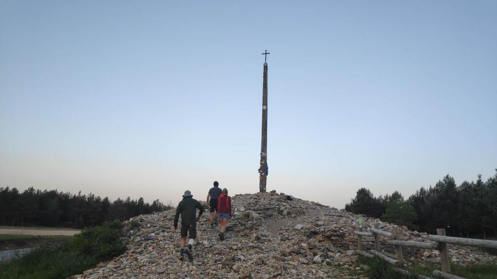 Tres peregrinos caminando hacia la Cruz de Hierro en el Camino de Santiago, rodeados de piedras y vegetación.