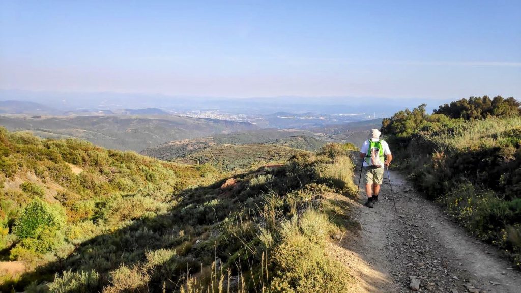 Un peregrino camina por un sendero montañoso con vistas panorámicas de un valle y montañas en la ruta Foncebadón-Ponferrada del Camino de Santiago.