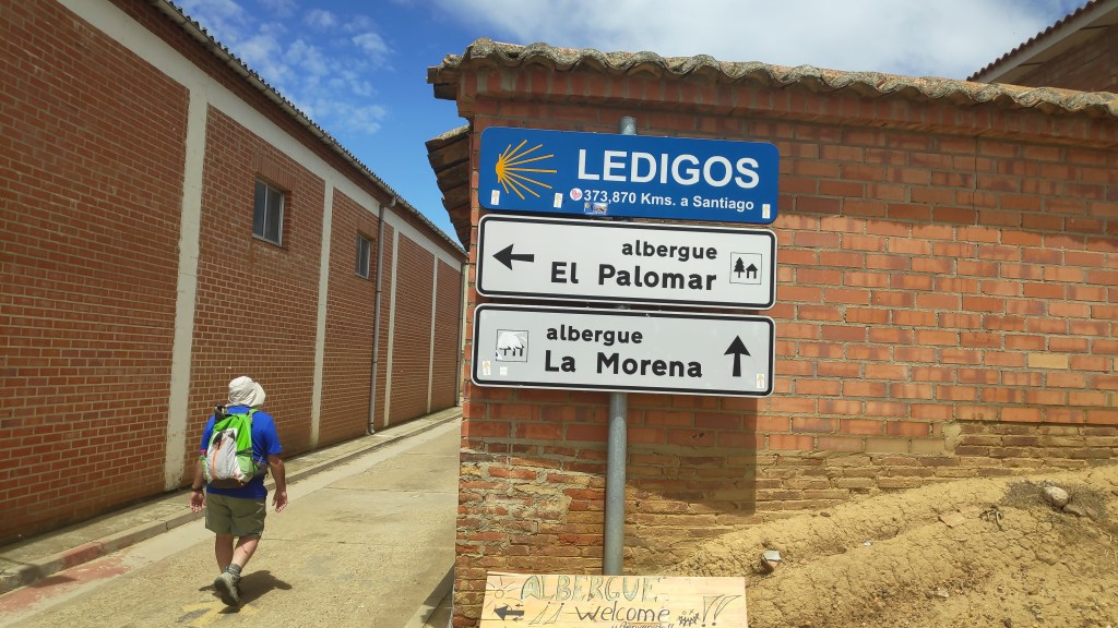 Etapa 17:Carrión de los Condes-Ledigos - Caminando por Mallorca Un peregrino caminando hacia Ledigos, con un cartel que muestra la distancia a Santiago y las direcciones de los albergues El Palomar y La Morena.