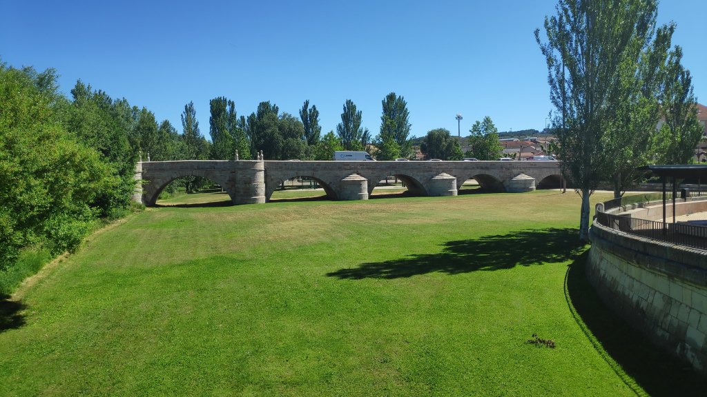 Puente de piedra de varios arcos sobre un campo verde, con árboles al fondo y un cielo despejado.