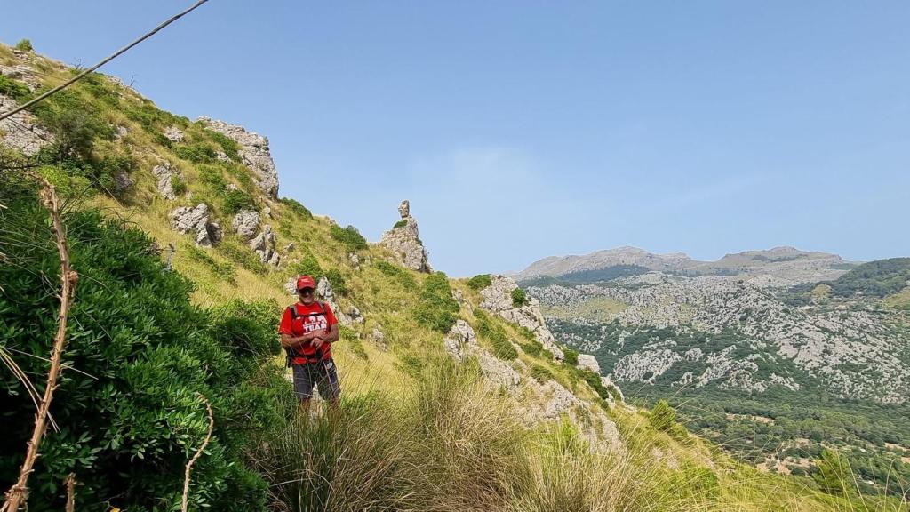 Senderista en la ruta 'Paso del Diablo' junto a una formación rocosa en un paisaje montañoso, con vegetación densa a su alrededor y montañas al fondo.