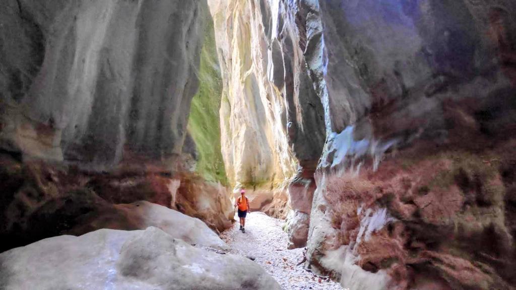 Caminante en el lecho de un cañón estrecho, rodeado de altas paredes de roca con tonos terrosos y verdes, en la ruta Torrente de Pareis.
