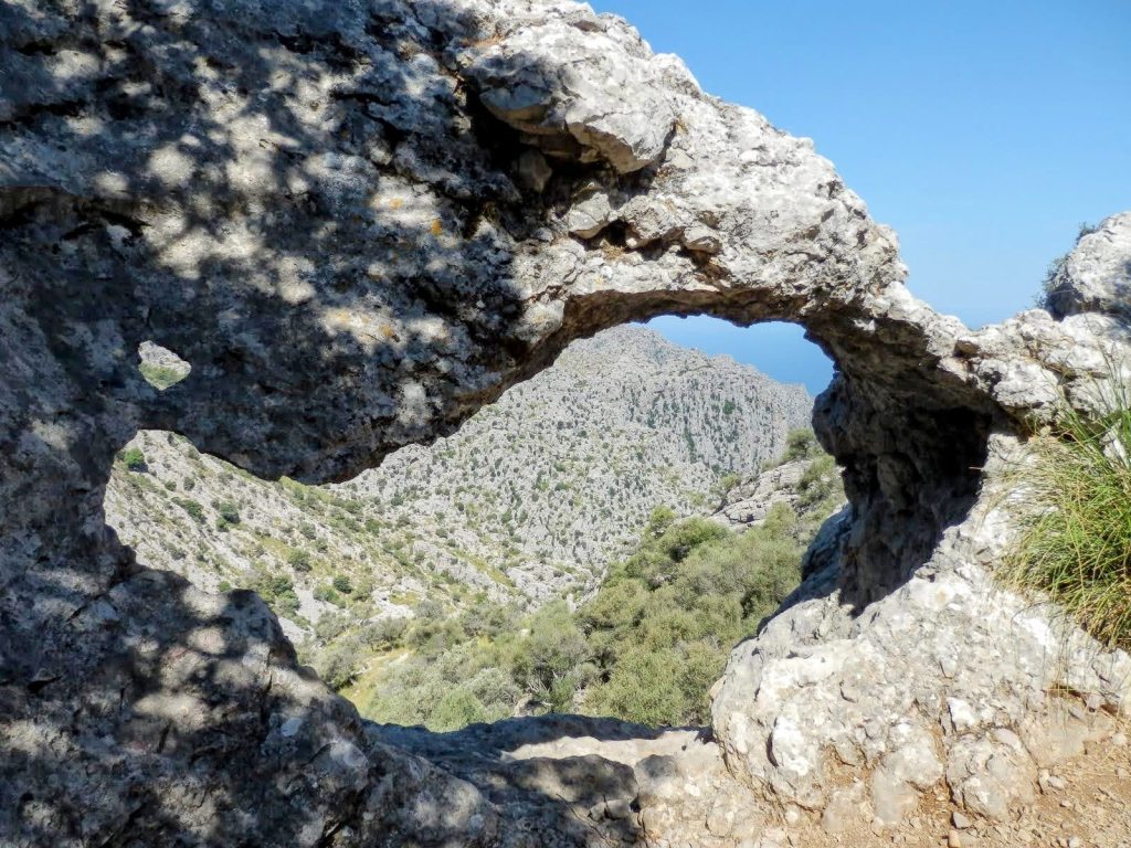 Vista a través de un arco natural de roca en la Serra de la Tramuntana, mostrando montañas cubiertas de vegetación y cielo azul al fondo.