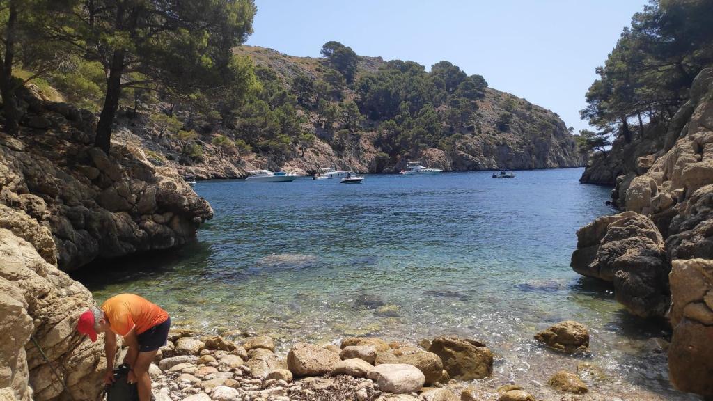 El Fumat y Cala en Gossalba - Caminando por Mallorca Vista de Cala en Gossalba, con un hombre al fondo preparando su equipo, rodeada de rocas y árboles, y con barcos en el agua.