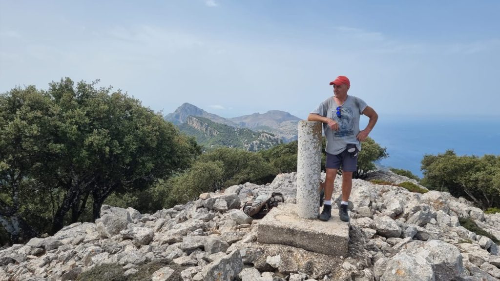 Hombre de pie junto a un vértice geodésico en la cima de la Mola de Planicia, rodeado de rocas y árboles, con vistas al paisaje montañoso y al mar en el fondo.