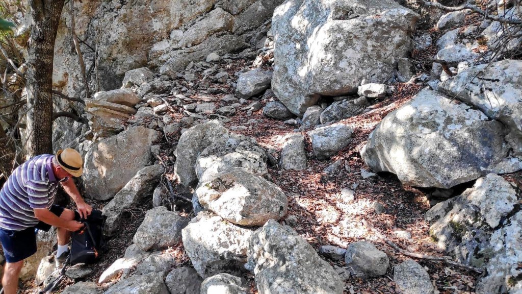Un sendero rocoso en una ruta de senderismo con piedras grandes y un árbol, mientras un hombre con un sombrero revisa su mochila.
